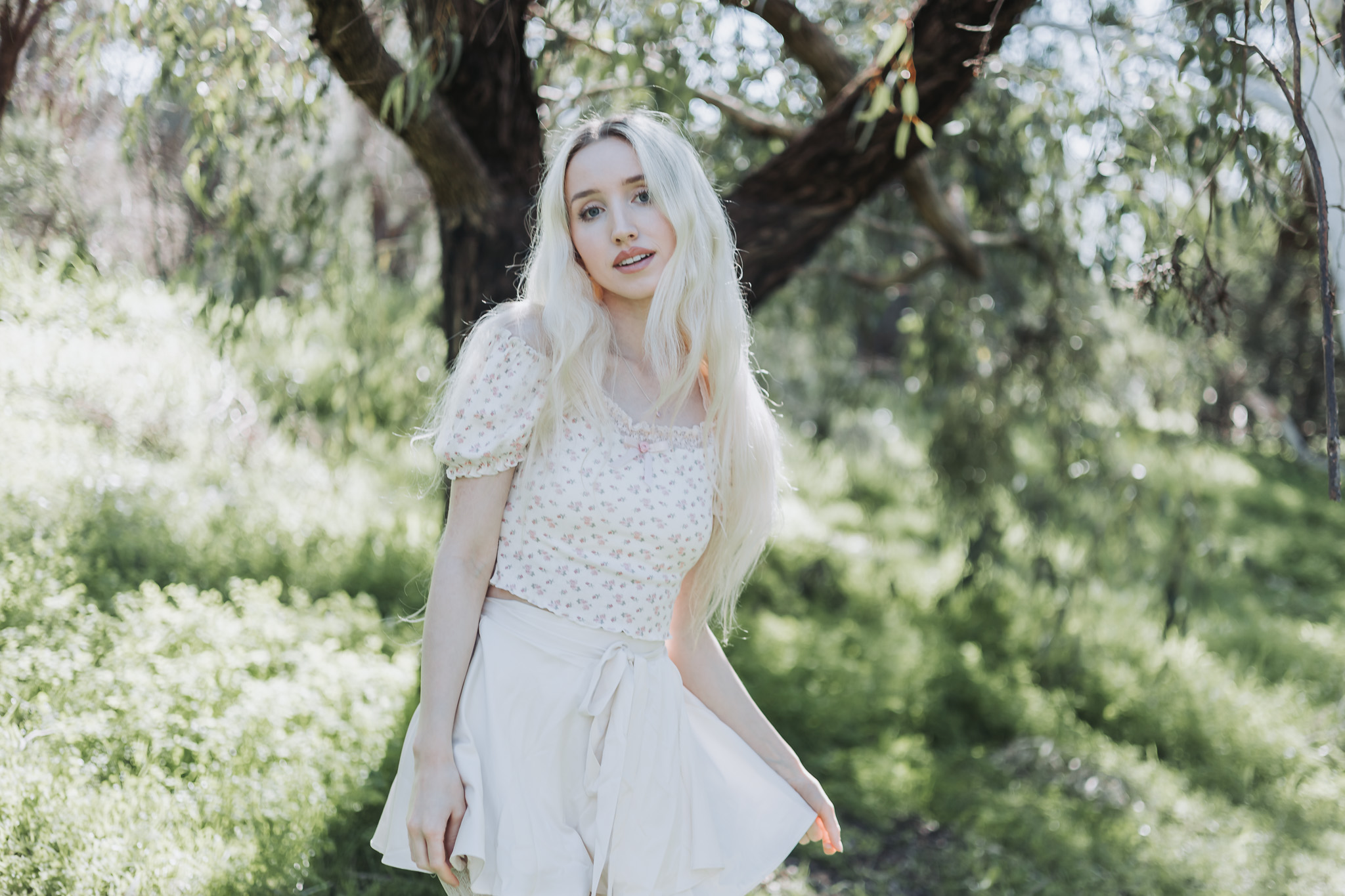 Woman with long blonde hair in a white floral dress posing among sunlit trees